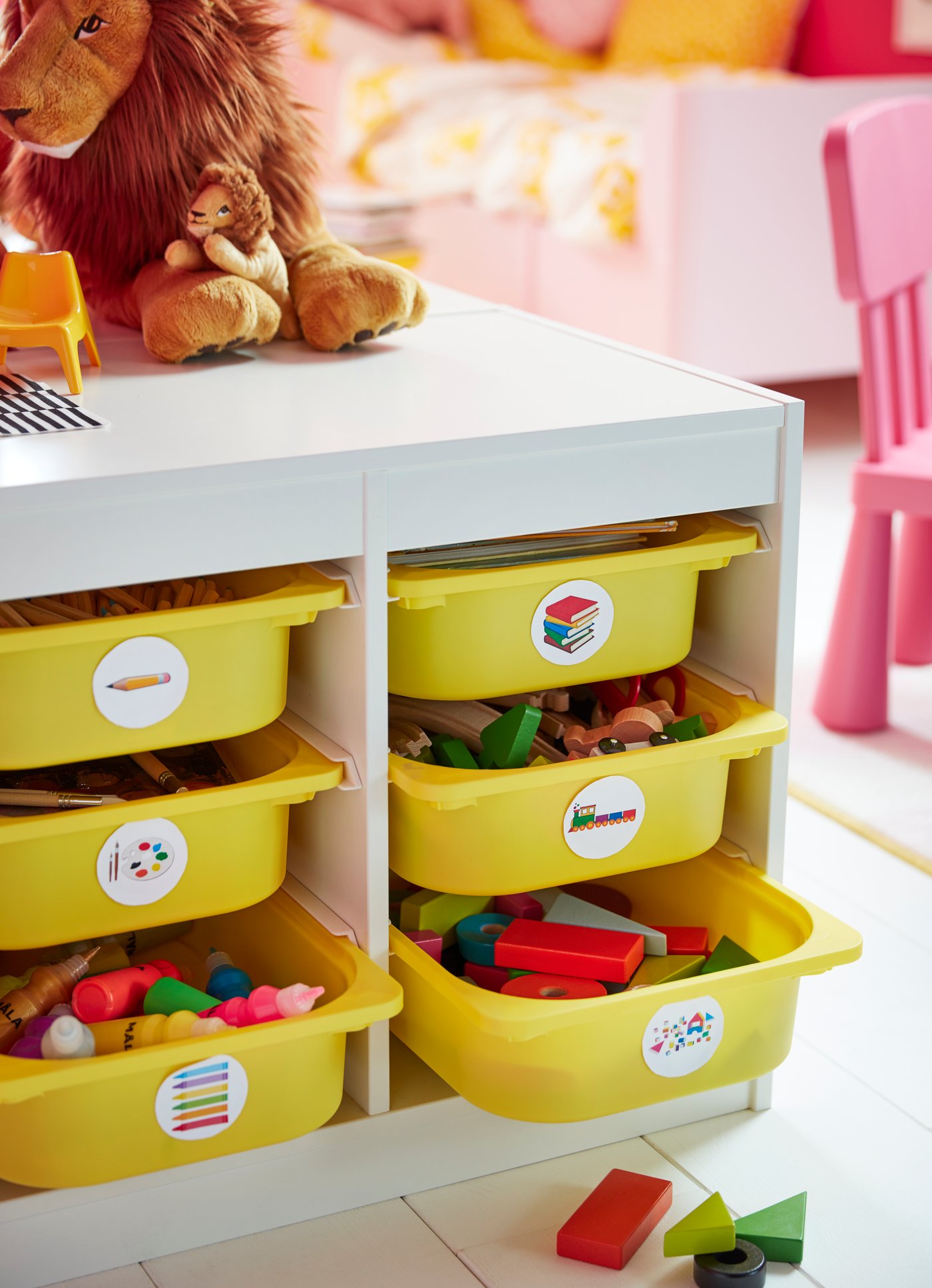 Un mueble TROFAST blanco con cajas amarillas en una pieza con muebles infantiles. Las cajas tienen etiquetas con dibujos que indican su contenido, y sobre el mueble hay dos peluches con forma de león. 