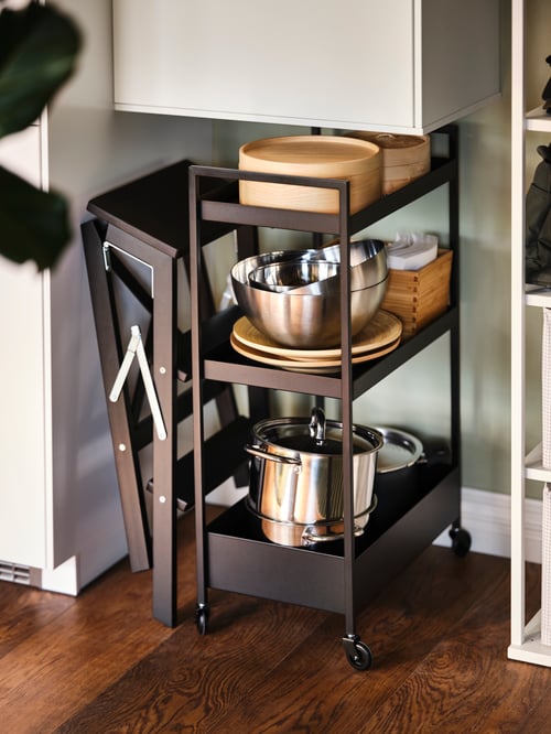 A folded black BEKVÄM stepladder with 3 steps against a wall, next to a black NISSAFORS trolley holding pots, pans and bowls.