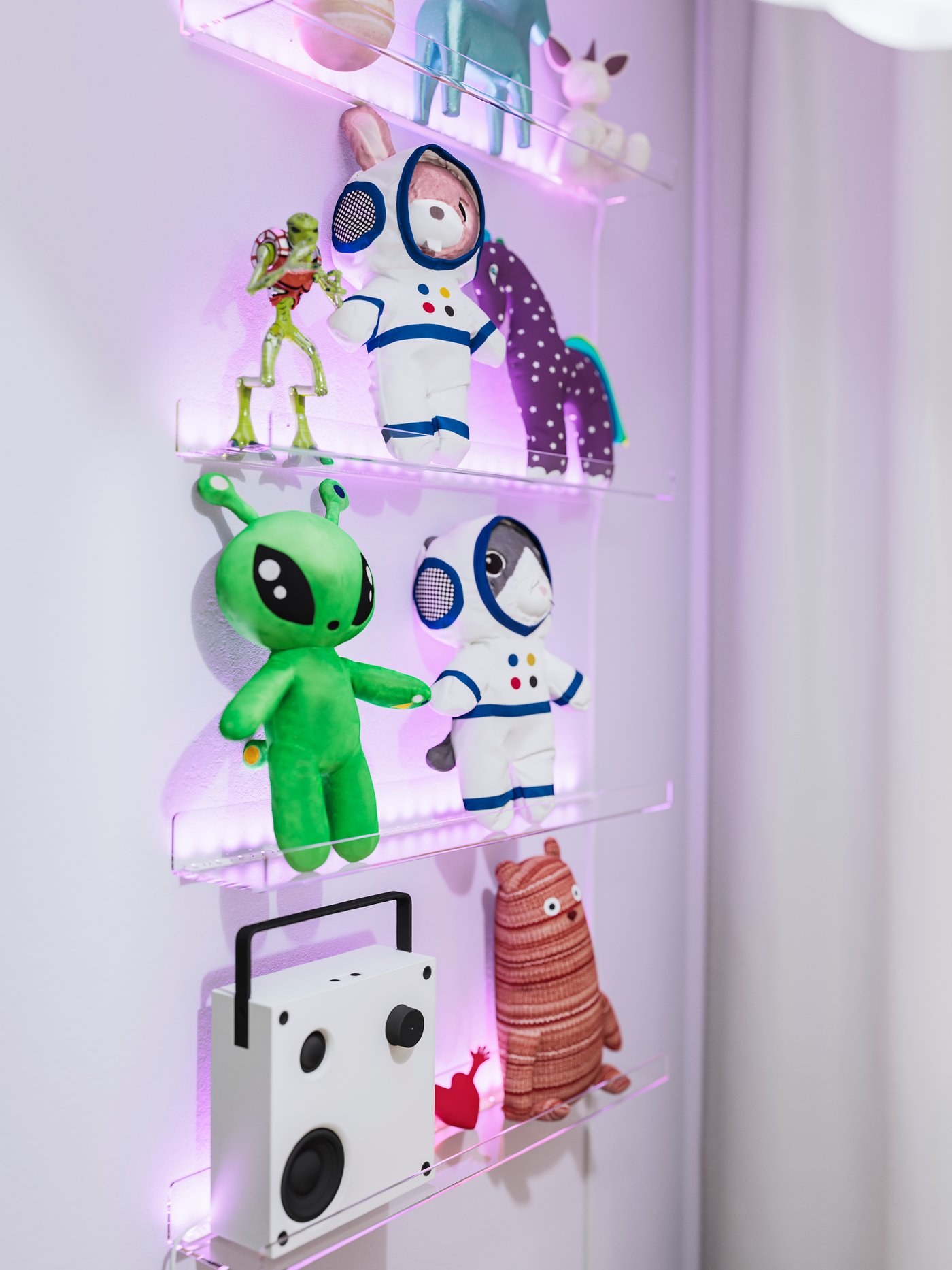 Vier transparente MELLÖSA Bilderleisten hängen an einer Wand. Auf ihnen befinden sich ein VAPPEBY Bluetooth-Lautsprecher und Spielsachen, unter anderem auch AFTONSPARV Stoffspielzeuge.