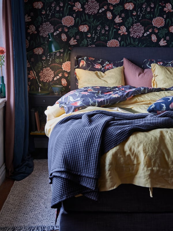 Prikaz kreveta s poplunom na cvjetni uzorak i sivim prekrivačem. Pored njega nalazi se noćni ormarić s lampom za čitanje.