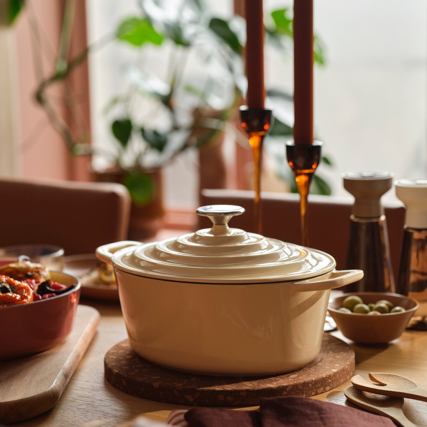 A VARDAGEN pot in enamelled cast iron in light yellow stands on a wooden dining table surrounded by food and candles.