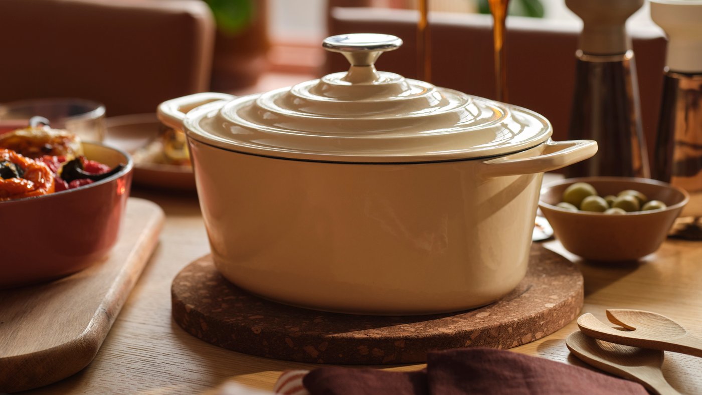 A VARDAGEN pot in enamelled cast iron in light yellow stands on a wooden dining table surrounded by food and candles.