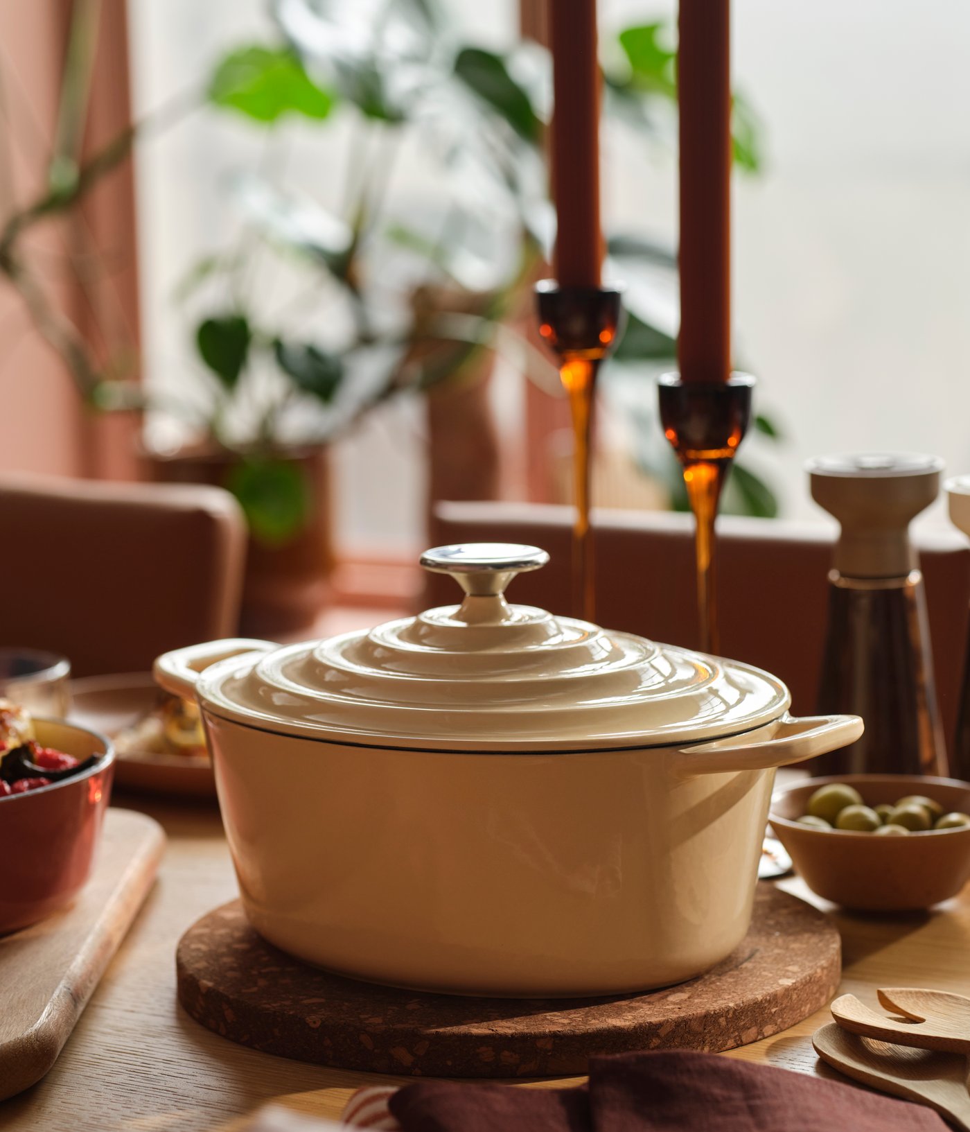 A VARDAGEN pot in enamelled cast iron in light yellow stands on a wooden dining table surrounded by food and candles.