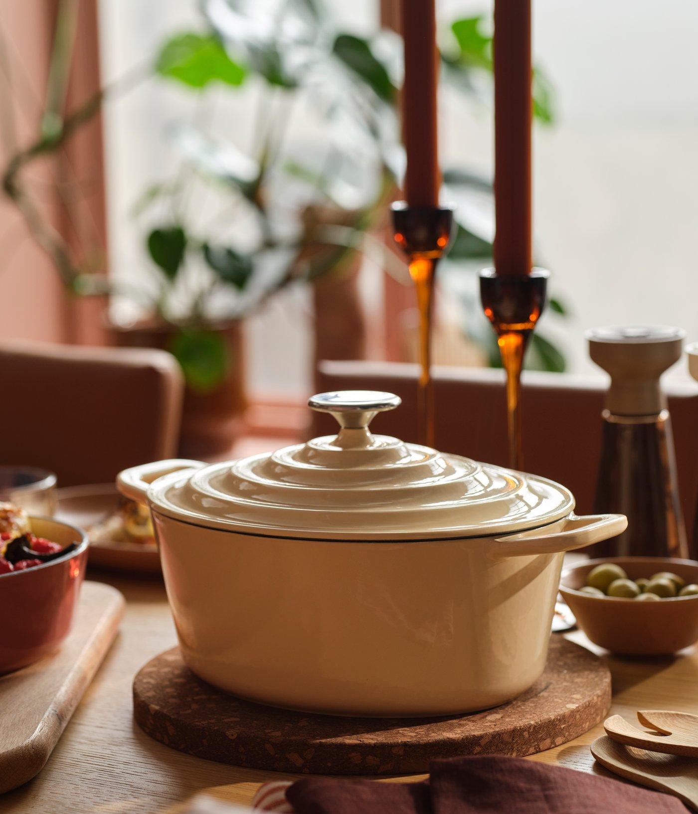A VARDAGEN pot in enamelled cast iron in light yellow stands on a wooden dining table surrounded by food and candles.
