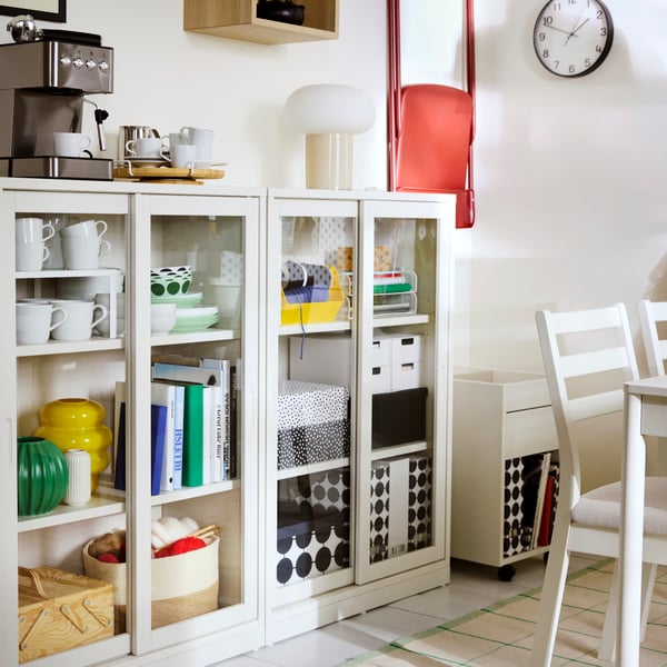 A room with two TONSTAD glass door cabinets filled with diverse items and a TONSTAD drawer unit on castors in the corner.
