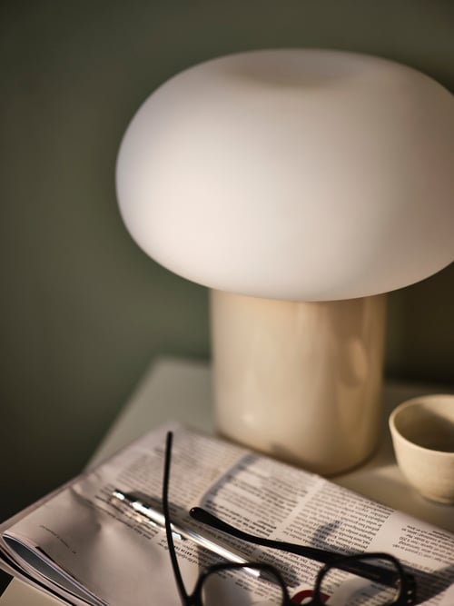 Une lampe de table DEJSA en verre opalin beige/blanc éteinte, sur une surface avec un journal, des stylos et une paire de lunettes.