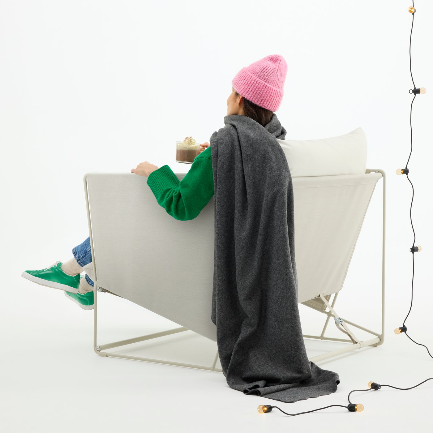 A person wearing a wool hat sits in an outdoor chair, draped in a grey DAGGKÅPOR throw.
