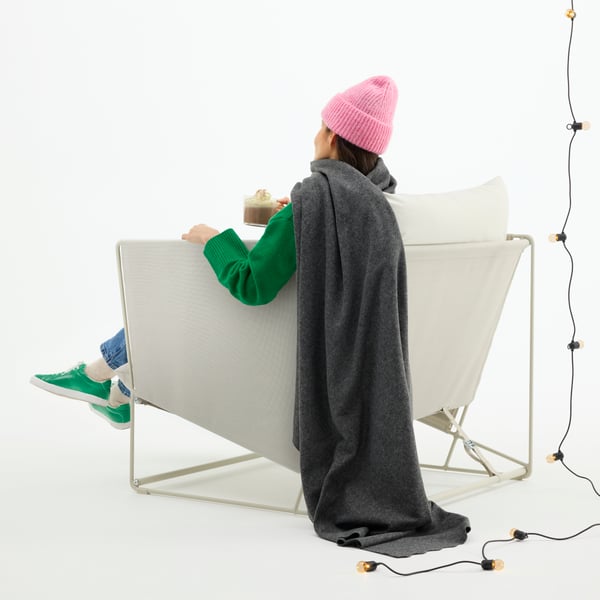 A woman sits in an outdoor chair, draped in a grey DAGGKÅPOR blanket. She’s wearing a wool hat, drinking coffee from a mug.