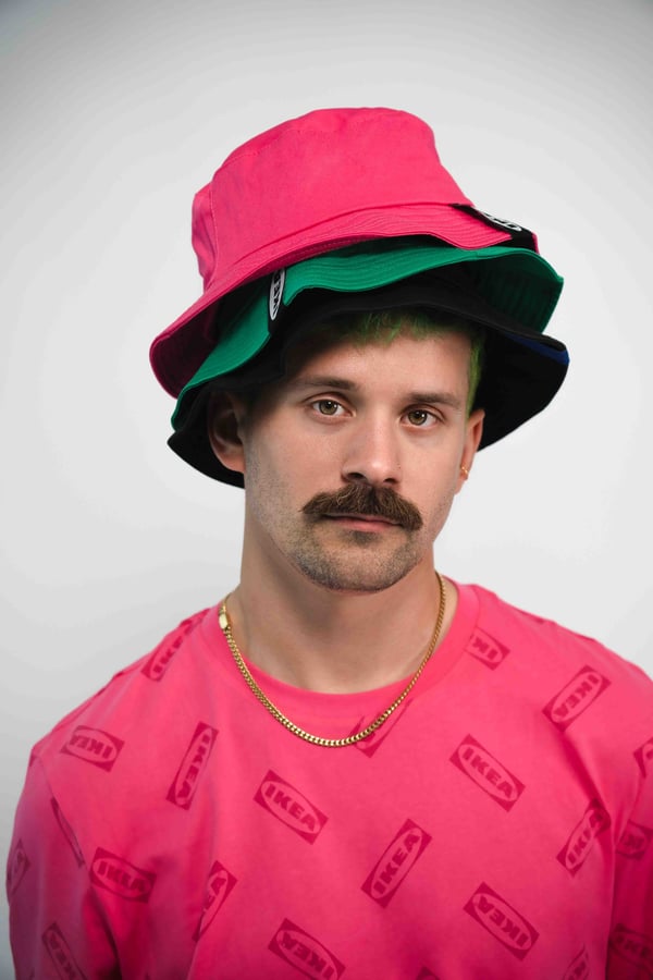 Modell bär två staplade bucket hats i rosa och grönt samt en rosa AURTIENDE t-shirt med IKEA-logotyp och ett guldfärgat halsband