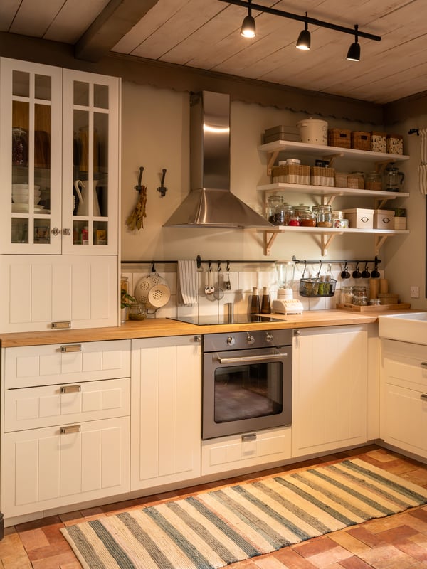 A well-lit, cozy kitchen interior with beige and white cabinetry. There is a stainless steel range hood above a stove, and to the right of the stove is an oven. Open shelves mounted on the wall hold various dishes and jars. On the left side, there’s a white cabinet with glass doors. The kitchen has several utensils hanging on the wall, and there’s a striped rug on the floor. The warm lighting and organized layout give this kitchen a welcoming and functional atmosphere.