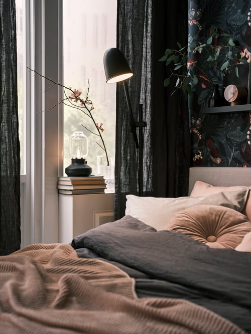 Bedroom setting featuring a grey table lamp, a bed with a grey duvet, cushions, and a throw, and a floor reading lamp.