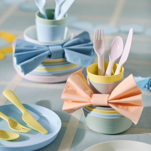 KALAS plates, bowls, cutlery and mugs in three piles on a light blue check tablecloth with pastel coloured bows around them.