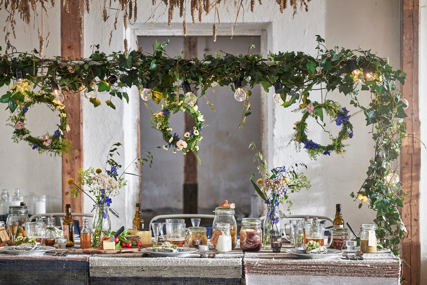 Rustikale Tafel mit Geschirr, Blumen und hängenden Grünpflanzen und Kränzen, dekoriert mit Lichtern unter Holzträgern in heller Umgebung.