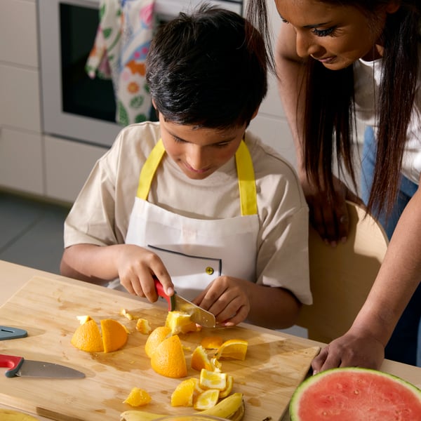 A little boy carefully cutting an orange with the help of SMÅBIT knife, the mother watching from the side.