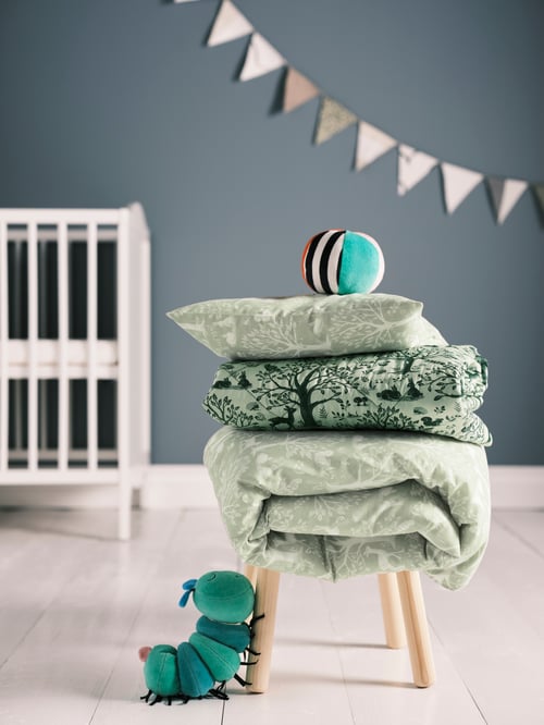 A folded quilted blanket and a folded duvet set is on a children's tool, with a green toy at the base and a cot in the back.