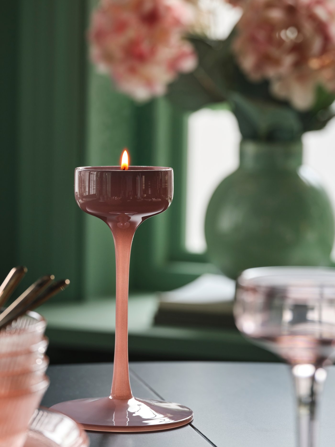 Ett tänt värmeljus i en elegant hög rosa ljushållare av glas på ett bord, med en grön vas och stilren heminredning i bakgrunden.