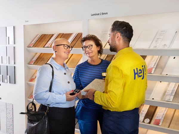 A smiling business client in an IKEA store stands in front of shelves with material samples, getting consulted by an interior designer and her co-worker.