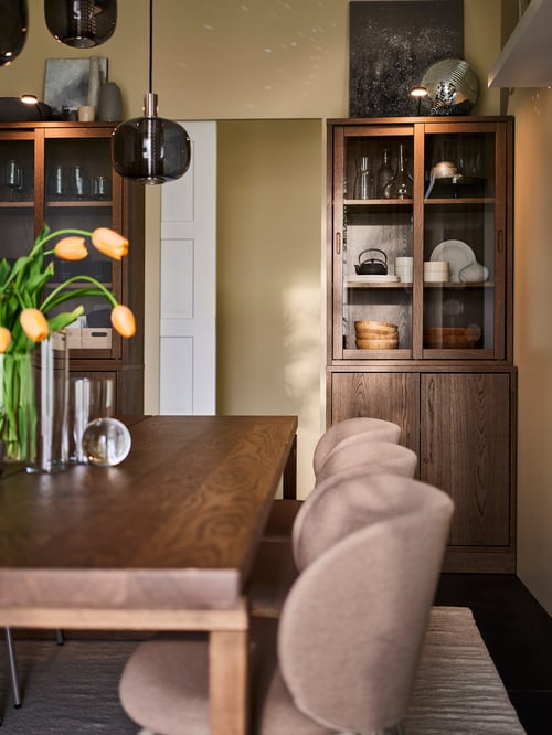 A living room with two TONSTAD storage combinatios with sliding glass doors and a cabinet lighting, a table and beige chairs.