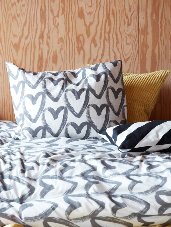 A bed with two cushions and a LYKTFIBBLA quilt cover and pillow case with gray hearts on a white background.