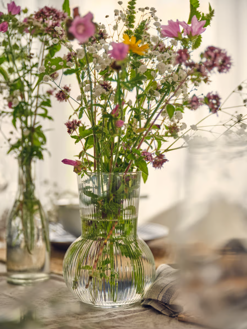 Glasvase mit bunten Wildblumen auf einem Tisch, im Hintergrund weitere Vasen mit ähnlichen Blumenarrangements.