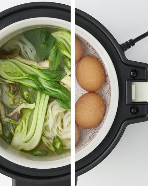 Un multicuiseur IKEA est ingénieusement représenté en train de préparer simultanément deux repas différents, avec une soupe verte vibrante contenant du bok choy et des nouilles d'un côté, et des œufs bruns parfaitement cuits à la vapeur de l'autre.