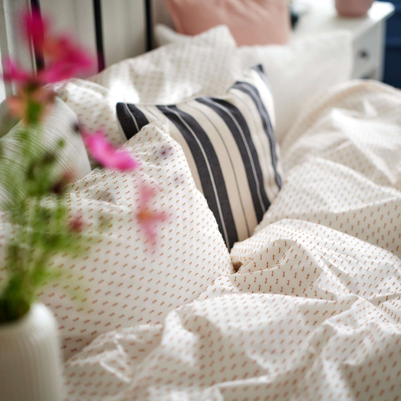 The top of a bed with white-beige BANDKNIPPE bed linen on it and cushions in various covers including KORALLBUSKE.