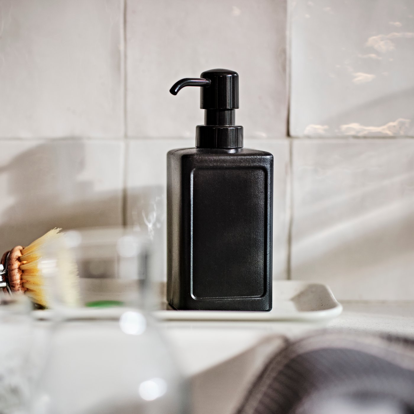 Siv RINNIG dozirnik za tekoče milo na majhnem belem pladnju na kuhinjskem pultu ob steni z belimi ploščicami.