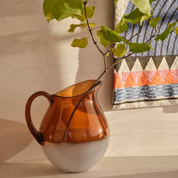 Oranssissa puoliksi läpinäkyvässä lasikannussa on puun oksa, jossa vihreät lehdet. Kannu on sijoitettu vaalealle puutasolle, jonka tausta on samaa puumateriaalia. Taustalla näkyy pieni osa värikkäästä seinäryijystä, jossa on erivärisiä raitoja ja kolmioita.