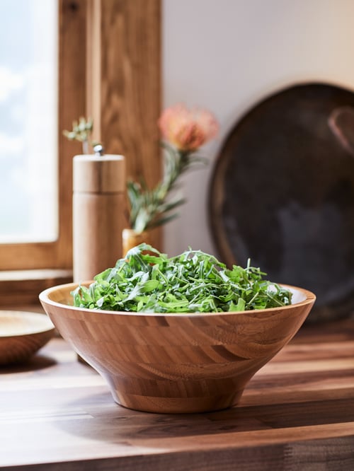 A wood-finish bamboo GRÖNSAKER serving bowl filled with green salad leaves on a wooden kitchen worktop.