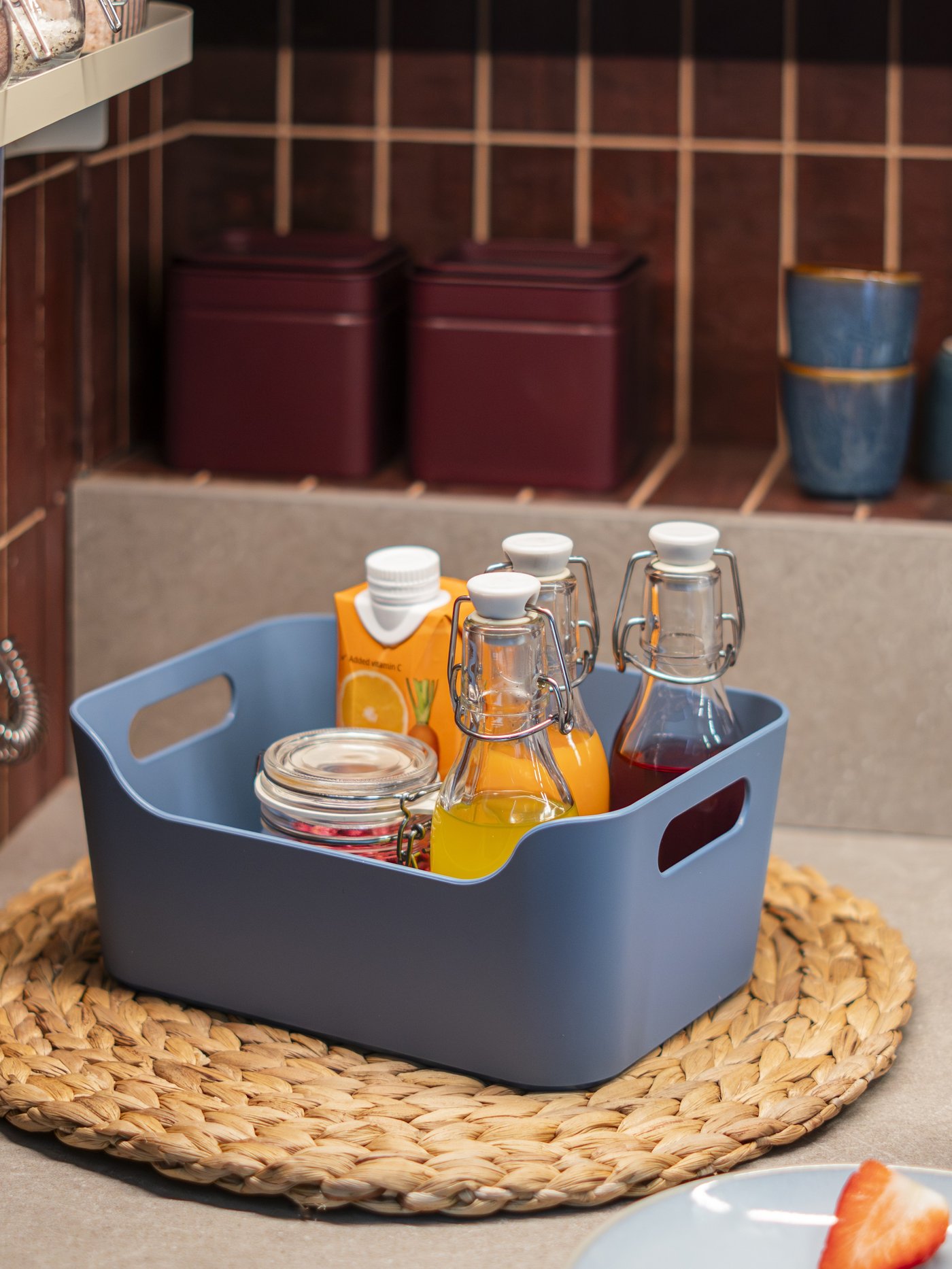 Tazza BLOMVECKLARE rosa chiaro appoggiata sul piano cucina, accanto al contenitore blu UPPDATERA con bottiglie e succhi di frutta.
