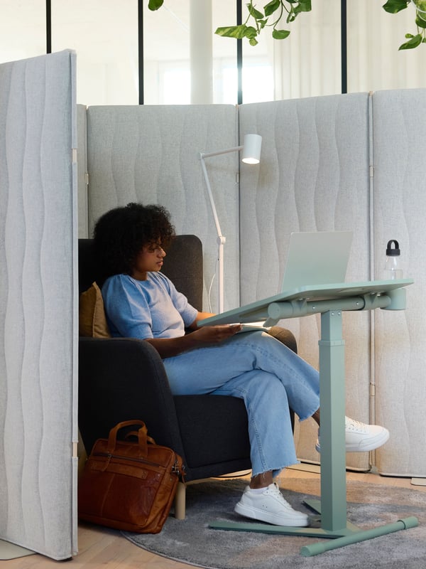 A person sits in a dark grey armchair by a green MITTZON laptop table with castors that is tilted down, holding a laptop.