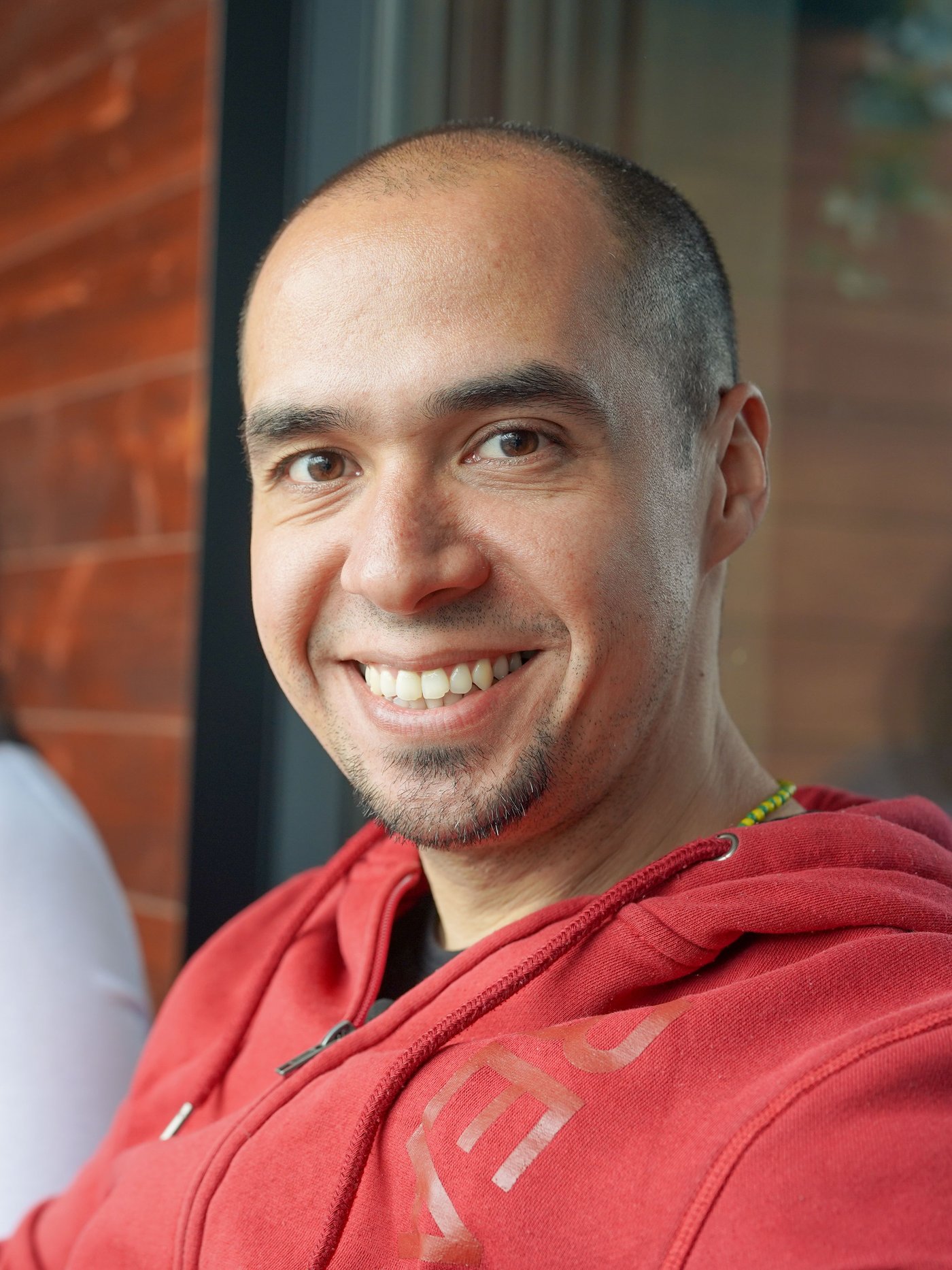 A man sits outdoors in his red shirt and smiles at the camera.