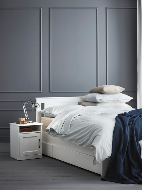 Bedroom with grey wall with a stack of pillows placed on a bed frame with storage boxes and bedspread, and a bedside table.