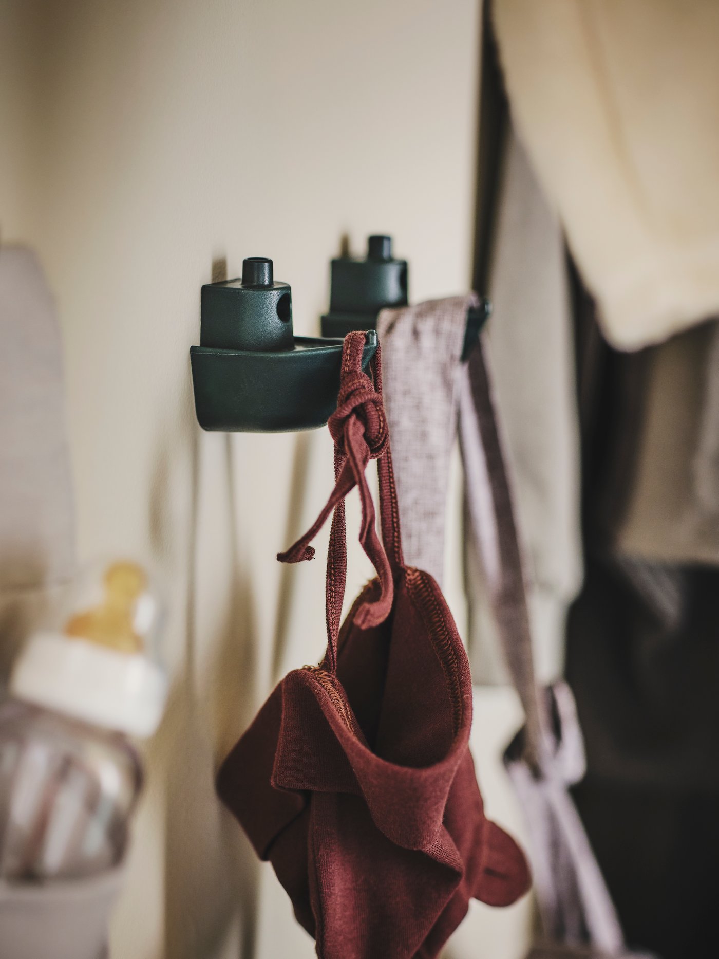 Dva zelené háčky KNOPARE jsou umístěné na béžové stěně. Na jednom visí rezavě hnědá čepice, na druhém je šedá taška.