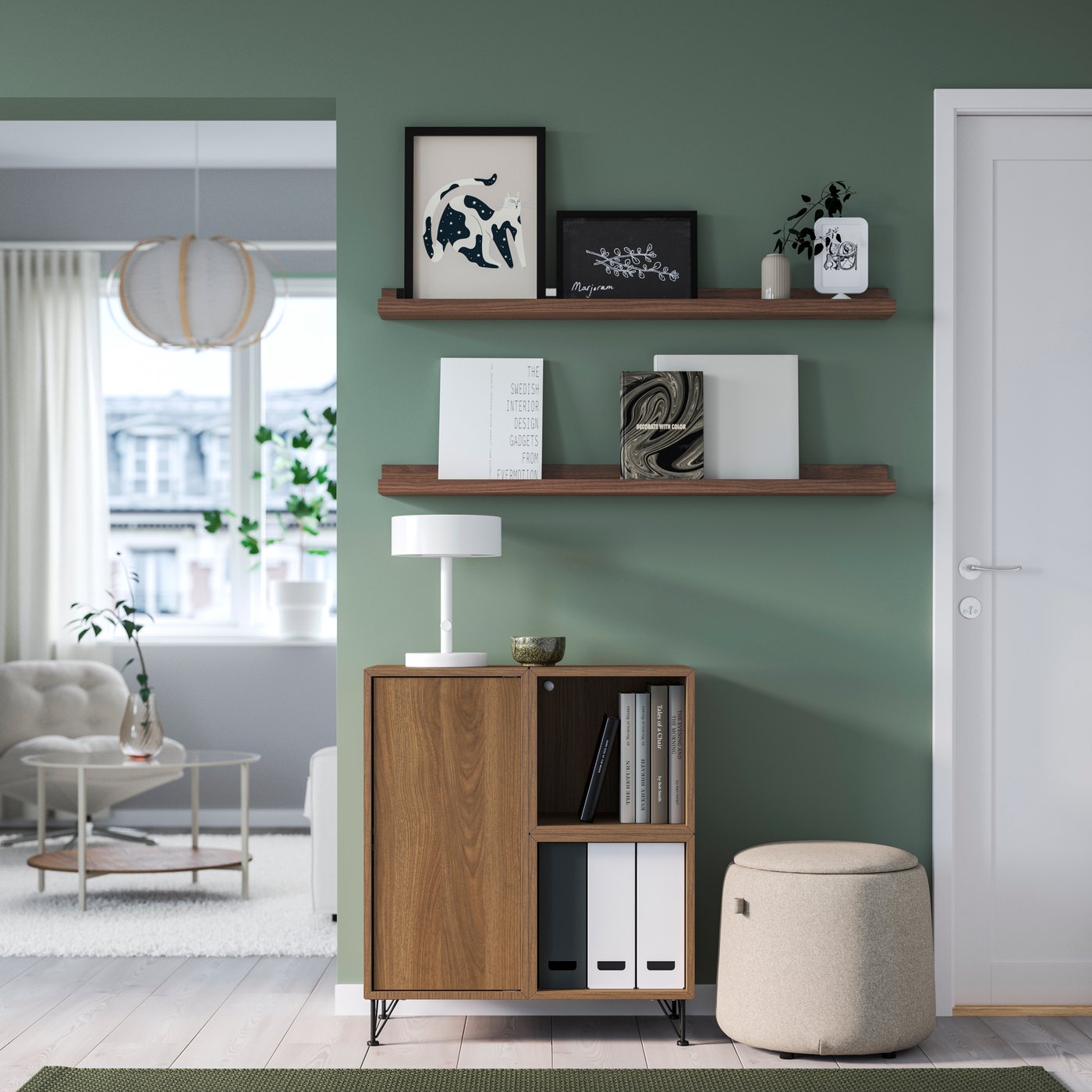 Ingresso moderno con mobile contenitore in legno, mensole MOSSLANDA alla parete con libri e quadri e pouf imbottito chiaro accanto alla porta.
