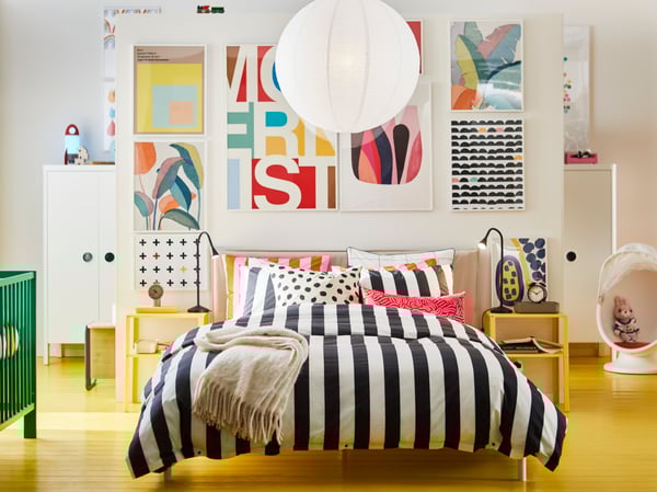 Furnished bedroom with yellow floor with the bed in the middle and a large picture wall behind. 