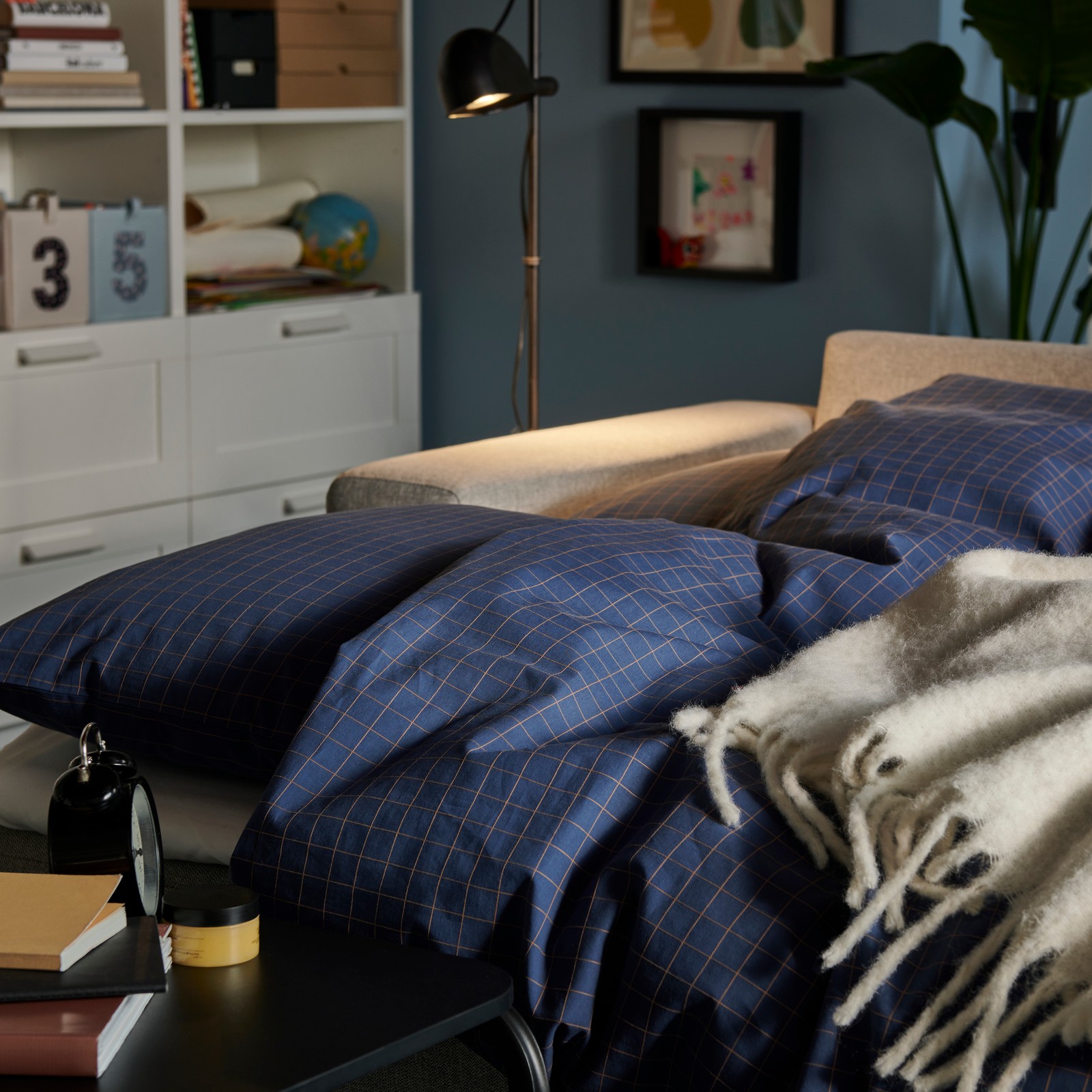 Unha sala de estar cunha luz tenue, un sofá cama BÅRSLÖV con roupa de cama DOFTAKLEJA e unha mesa auxiliar OLSERÖD xunto ao sofá.
