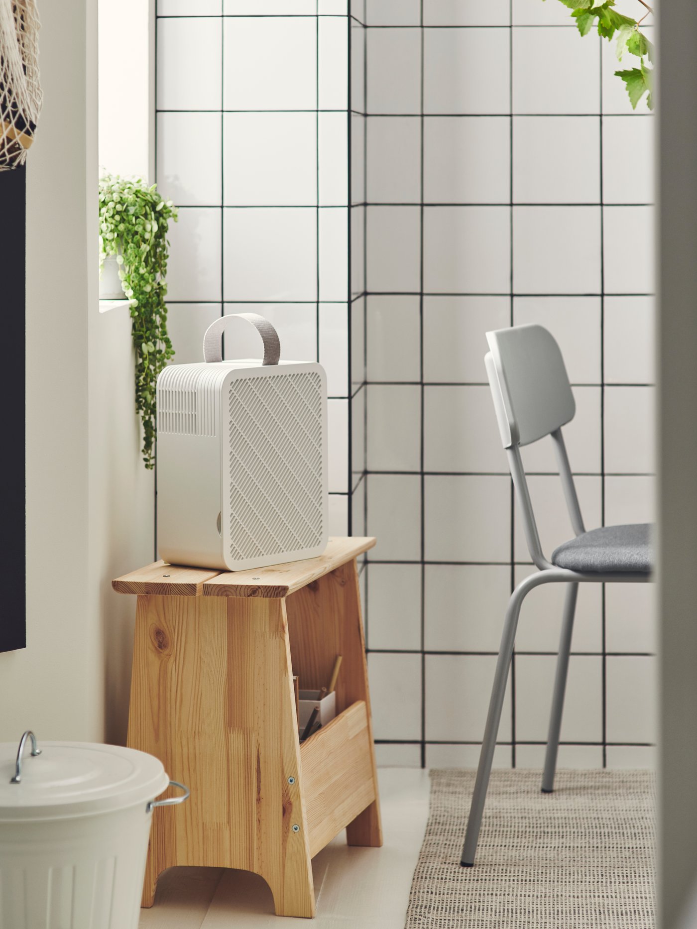 Ambiente de baño con un purificador de aire UPPÅTVIND colocado sobre un taburete PERJOHAN con almacenaje. El taburete está junto a una silla gris.