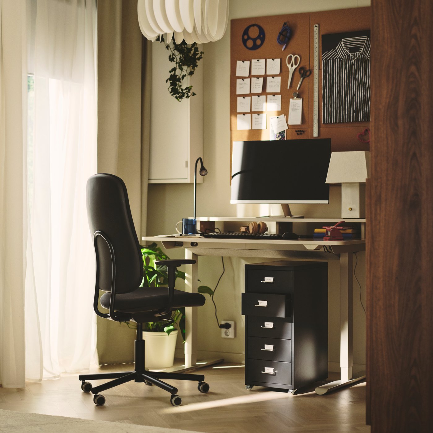 A home office with a beige TRAVSPORT desk sit/stand next to a black rolling chair and potted plants.