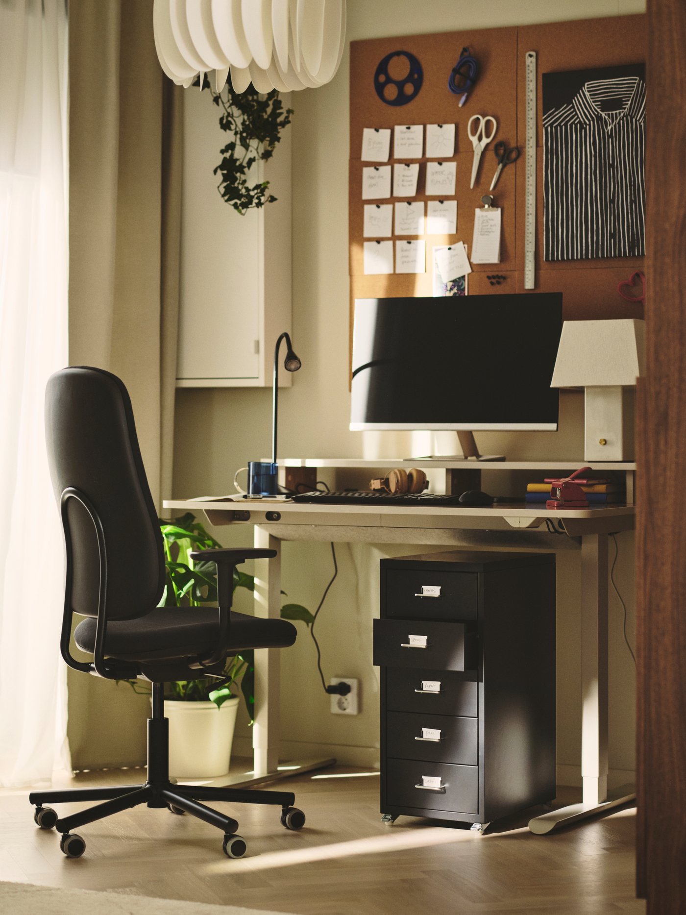 Home office con scrivania regolabile TRAVSPORT beige, sedia girevole nera, cassettiera e piante in vaso accanto a una grande finestra luminosa.