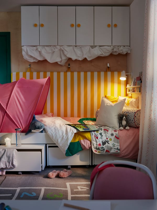 A child’s room with a SLÄKT bed and storage boxes with a FUBBLA wall lamp at one end, and a SUFFLETT bed tent at the other.