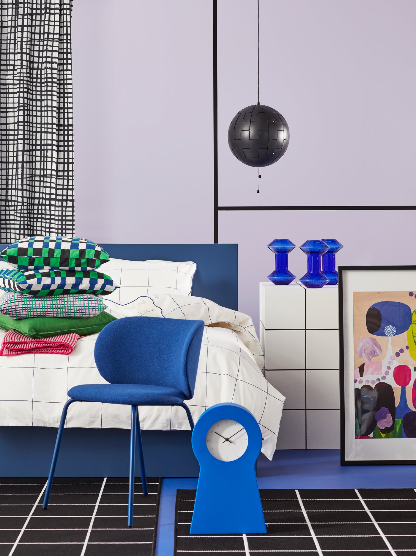 V popředí místnosti se nacházejí modré hodiny na černo- bílé podlaze. V pozadí místnosti je sedačka s polštáři a obraz.
