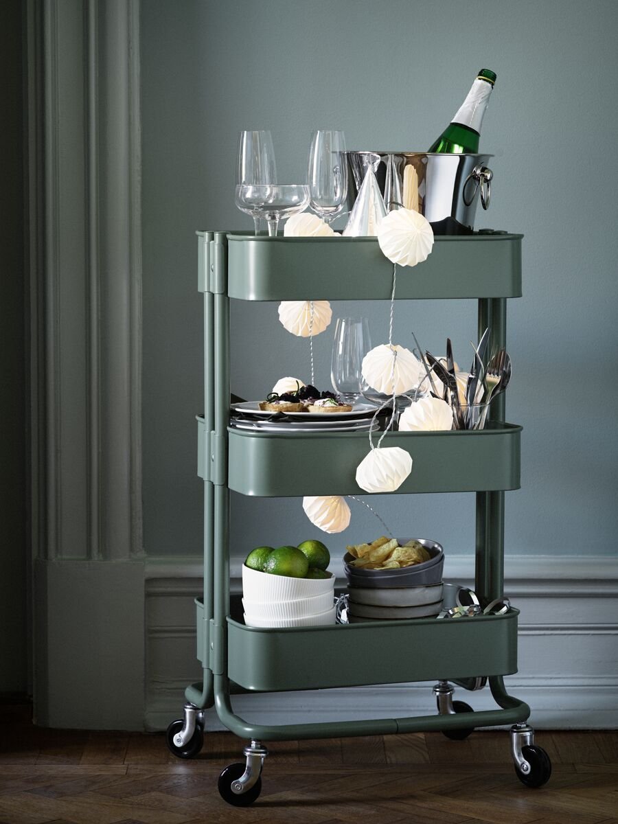 A green RÅSKOG trolley is packed with things for a party, including glasses, dinnerware and drinks to share.