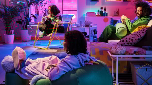 Ett modernt vardagsrum med färgstark belysning i rosa och lila toner. Tre personer sitter avslappnat: en i en gul stol och läser, en på en grön sittsäck, och en på en soffa med mönstrade kuddar. Rummet är inrett med gröna växter, hyllor med dekorationer, en randig matta och ett glasbord med små föremål. Stämningen är lekfull och mysig, med en kombination av naturligt ljus från fönstret och färgad belysning som skapar en kreativ atmosfär.