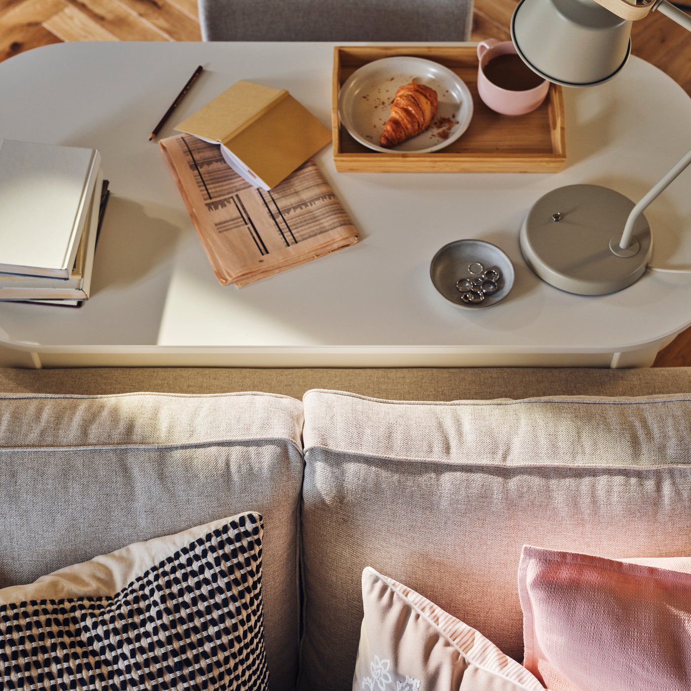 A tray with food and coffee, a lamp, a book and a newspaper stand on a ROSENTORP desk behind an EKHOLMA 2-seat sofa bed.