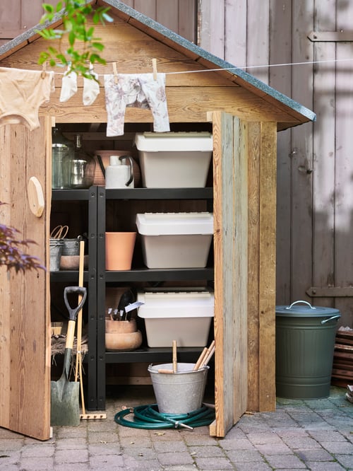 A BROR shelving unit inside a small shed holding gardening tools, with a washing line holding baby clothes in front of it.