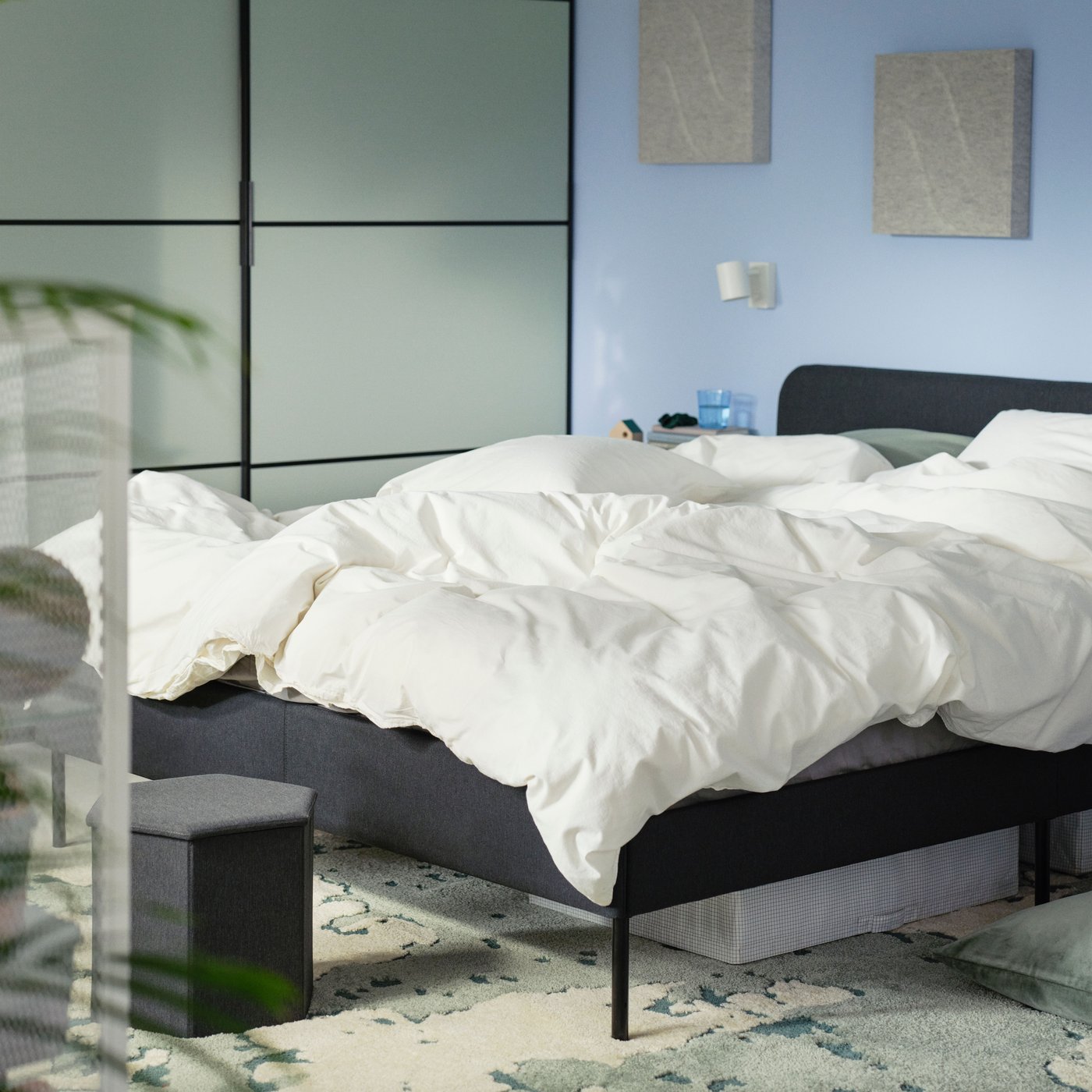 Ein grün/elfenbeinfarbener RINGKLOCKA-Teppich liegt unter einem schwarzen Bett mit weißer Bettwäsche.