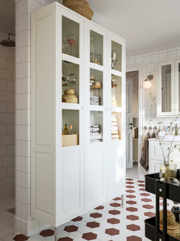 Three white TÄNNFORSEN high cabinets with doors, on two TÄNNFORSEN stainless steel legs, in a white tiled bathroom. 