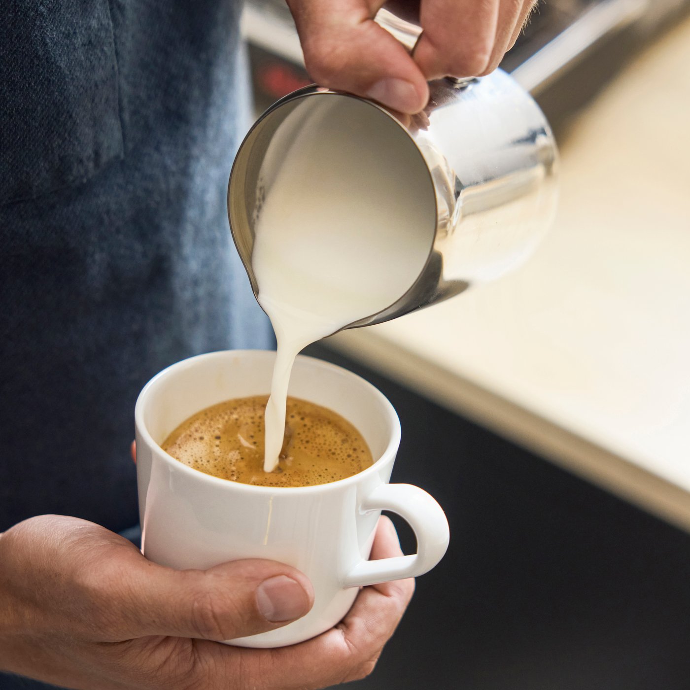 Mjölk hälls från en liten kanna i en kaffekopp, vilket skapar skum, med händer som håller kopp och kanna i kafémiljö.