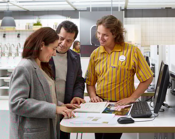 Et par får hjælp af en IKEA medarbejder mens de kigger i en brochure som ligger på et bord i en køkkenafdeling.